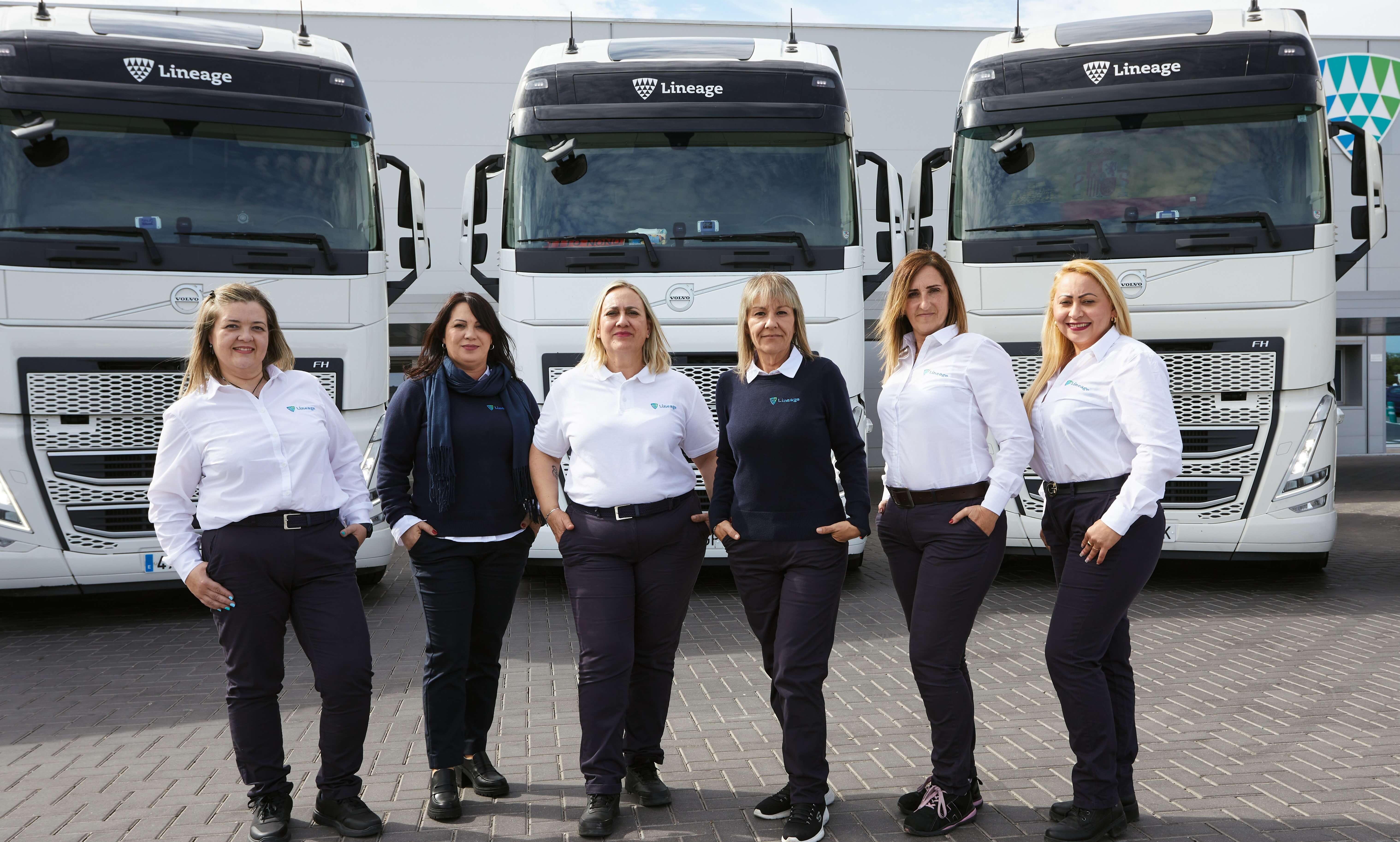 Five women drivers in white shirts and dark pants standing confidently in front of Lineage logistics trucks.