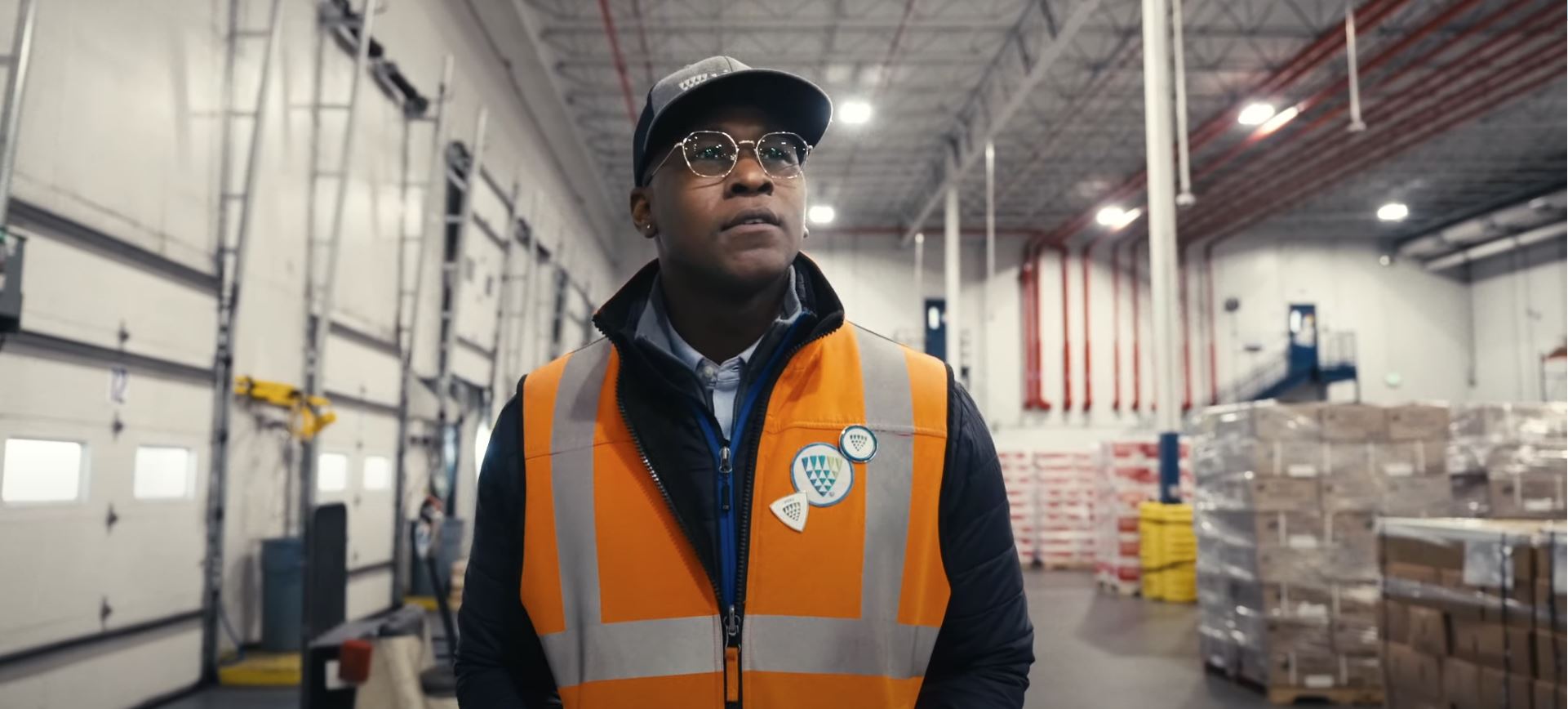 An attentive warehouse team member wearing a reflective orange vest and glasses walks through a spacious warehouse.