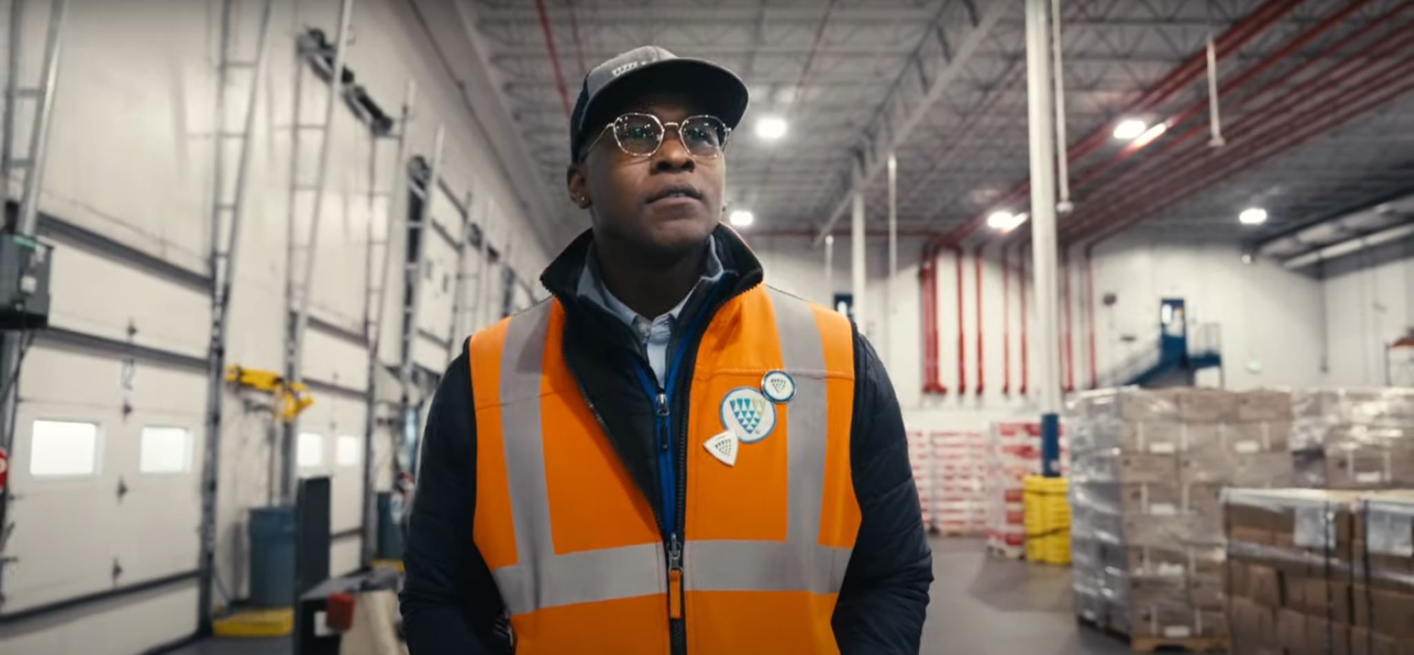 Alt text: "A focused individual, Brandon Richardson, donned in a high-visibility orange safety vest adorned with Lineage Logistics badges, stands proudly inside a vast warehouse. His attire, complete with a smart button-up shirt and stylish glasses, suggests a blend of professionalism and preparedness as he embarks on his journey through the Lineage Management Program. The background blur underscores the bustling environment of the warehouse, indicative of the dynamic workplace Lineage offers.
