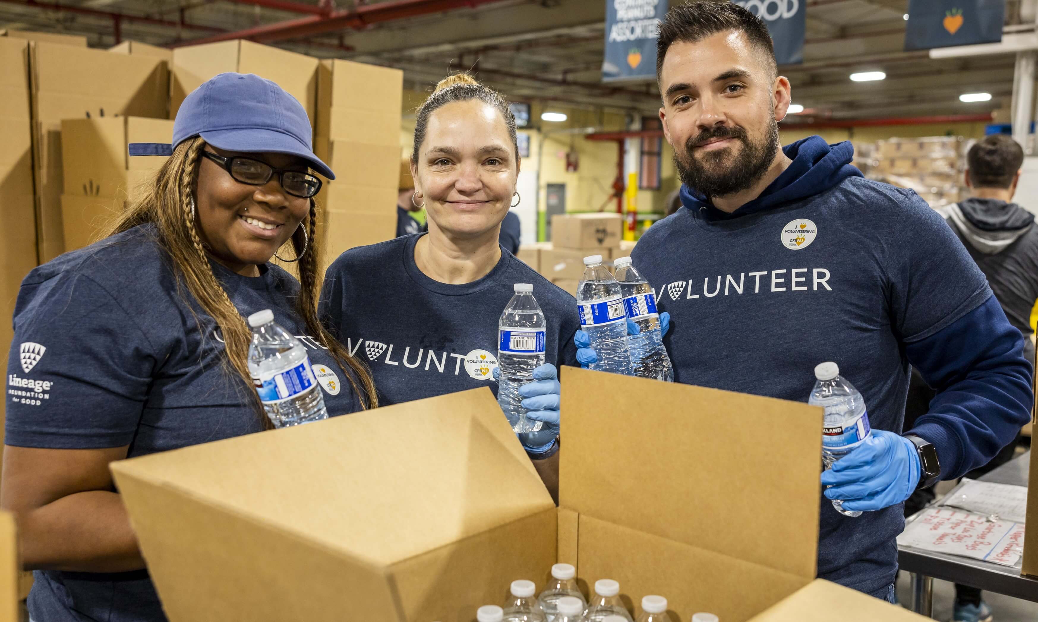 Three volunteers packing water bottles for distribution, wearing blue 'VOLUNTEER' t-shirts and smiling.