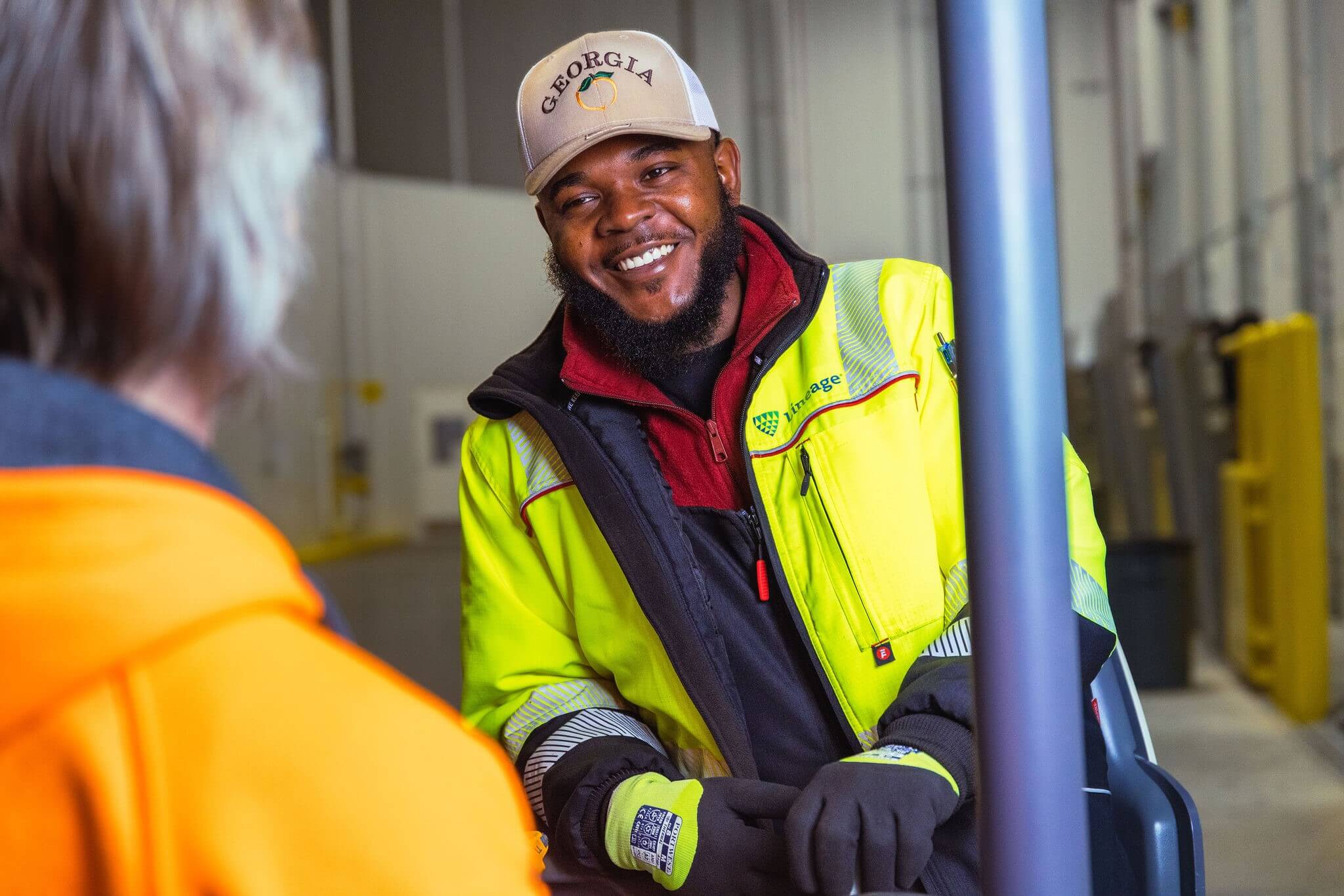 A smiling worker in a high-visibility jacket and 'Georgia' cap talking to a colleague in a warehouse.